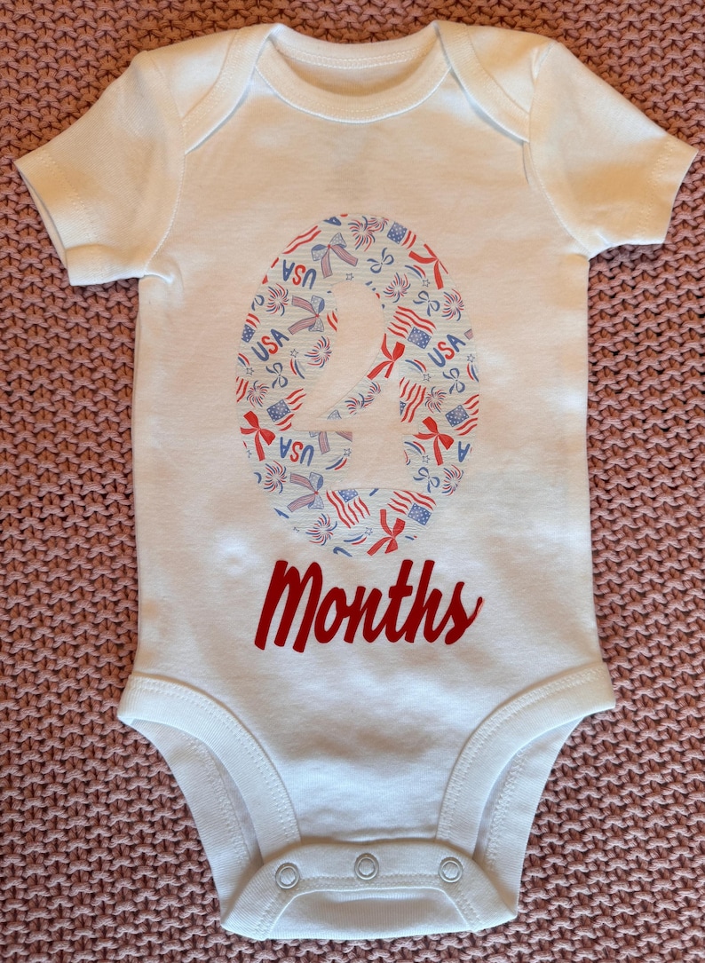 Może przedstawiać: Białe body dla dziecka z patriotycznym wzorem. Numer "2" wypełniony jest grafiką w kolorach czerwonym, białym i niebieskim w stylu USA. Słowo "Months" jest napisane czerwoną czcionką poniżej. Ubranko dla dziecka.
