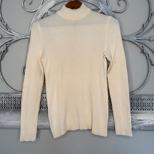 May include: Cream-colored turtleneck sweater with long sleeves. The ribbed knit sweater is displayed on a wooden hanger against a decorative background. The sweater's neutral color makes it a versatile wardrobe staple.