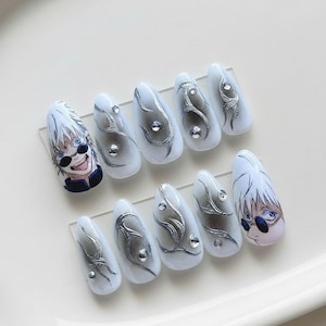 Peut inclure: Un ensemble de dix faux ongles blancs et argentés. Chaque ongle présente un design unique, comprenant des volutes argentées, des strass et des illustrations de personnages d'anime. Les ongles sont disposés sur une feuille de plastique transparente.