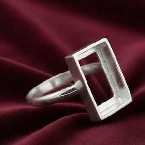 Puede incluir: Un anillo plateado con un engaste rectangular. El anillo se encuentra sobre un fondo de tela rojo oscuro, resaltando el diseño geométrico del anillo. La banda del anillo es lisa y redondeada.