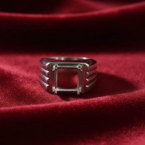 Könnte beinhalten: Ein silberfarbener Ring mit quadratischer Fassung für einen Edelstein. Der Ring hat ein dickes Band mit horizontalen Rillen und abgewinkelten Details. Die Fassung hat vier Krappen, um den Stein zu halten.