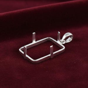 May include: A silver pendant setting with a rectangular frame and four prongs. The setting has a loop for a chain and is displayed against a dark red background. This jewelry component is designed for custom gemstone or crystal placement.