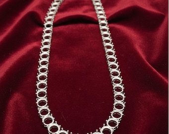 May include: A silver-colored necklace with multiple open circular settings, likely for gemstones, is displayed against a deep red velvet background. The necklace is designed to be worn around the neck.