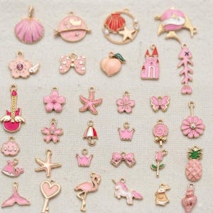 Könnte beinhalten: Eine Sammlung kleiner Metallanhänger in verschiedenen Formen, darunter Muscheln, Blumen, Sterne, ein Schloss, eine Gitarre, ein Flamingo, ein Einhorn, eine Ananas und Schleifen. Die Anhänger sind überwiegend rosa und haben Emaille-Details.