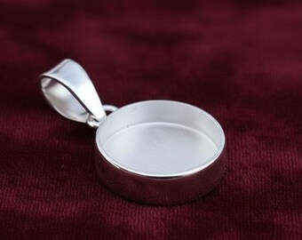 May include: A shiny silver pendant setting with a round, recessed area for a gemstone or other insert. The pendant has a loop for a chain and is set against a dark red background.