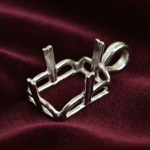 Könnte beinhalten: Ein silberner Anhänger mit rechteckiger Form und vier Krappen zur Aufnahme eines Edelsteins. Der Anhänger hat eine Schlaufe für eine Kette und liegt auf einem bordeauxfarbenen Stoff.