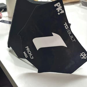 May include: Black cardstock with white text and graphics. The cardstock features the text "PK!", "TG|BUILT", "PRICKLY MSD", and a large white arrow. The cardstock is folded, possibly a product display.