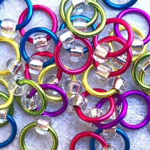 May include: A close-up of colorful metal rings with clear beads. The rings are in various colors, including green, blue, purple, and red. The beads have a silver and gold glitter design.