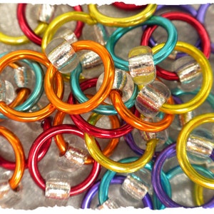 May include: A close-up of colorful metal rings in various colors, including red, orange, yellow, teal, and purple. Each ring is connected to a clear, cylindrical bead with a metallic interior. The rings are scattered, creating a vibrant and textured pattern.
