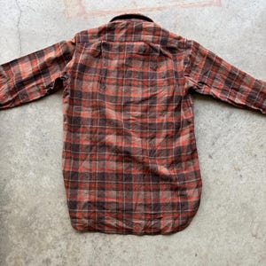 May include: A long-sleeve button-down shirt featuring a plaid pattern in shades of dark brown, tan, and vibrant orange. The shirt has a standard collar, a straight back yoke, and a rounded hemline, displaying a classic woven textile construction.