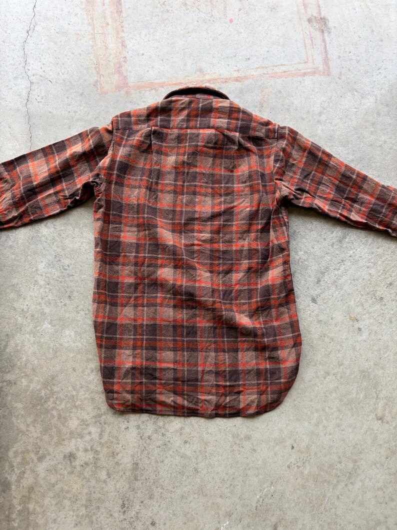 May include: A long-sleeve button-down shirt featuring a plaid pattern in shades of dark brown, tan, and vibrant orange. The shirt has a standard collar, a straight back yoke, and a rounded hemline, displaying a classic woven textile construction.