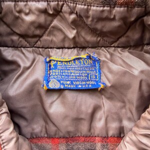May include: A brown and orange plaid jacket with a quilted brown lining. A blue rectangular Pendleton label is stitched to the interior, featuring the text Pendleton, Pendleton Woolen Mills, Portland Oregon, 100% Virgin Wool, 15, Pure Virgin Wool, Made in USA.
