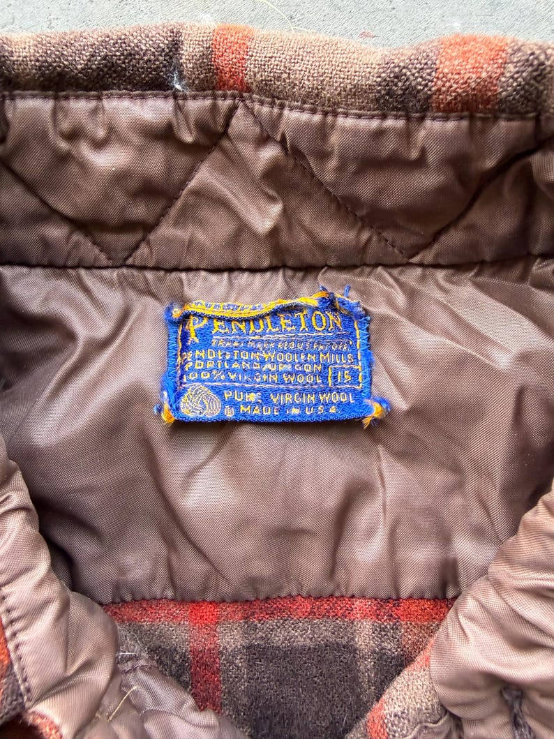 May include: A brown and orange plaid jacket with a quilted brown lining. A blue rectangular Pendleton label is stitched to the interior, featuring the text Pendleton, Pendleton Woolen Mills, Portland Oregon, 100% Virgin Wool, 15, Pure Virgin Wool, Made in USA.