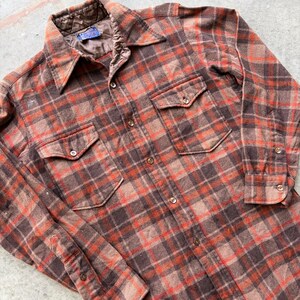 May include: A long-sleeve button-down shirt featuring a brown and orange plaid pattern. The shirt includes a pointed collar, two chest pockets with button closures, and a quilted brown lining visible at the neckline. The front placket has buttons.