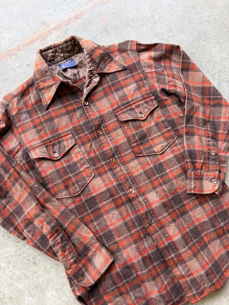 May include: A long-sleeve button-down shirt featuring a brown and orange plaid pattern. The shirt includes a pointed collar, two chest pockets with button closures, and a quilted brown lining visible at the neckline. The front placket has buttons.