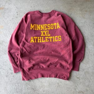 Könnte beinhalten: Ein kastanienbraunes Langarm-Sweatshirt mit Rundhalsausschnitt und dem gelben Schriftzug MINNESOTA XXL ATHLETICS auf der Brust. Das Kleidungsstück verfügt über gerippte Bündchen an Kragen, Ärmeln und Saum sowie eine klassische, sportliche Passform.