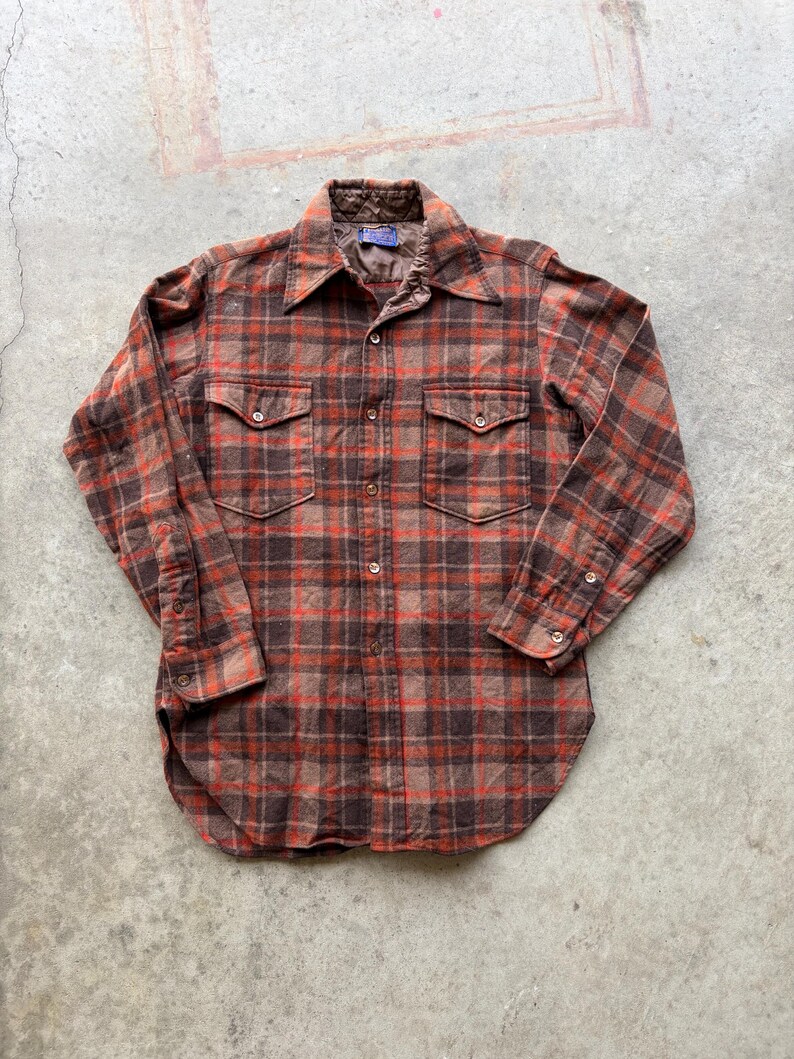 May include: A long-sleeve button-down shirt featuring a brown and orange plaid pattern. The shirt includes a pointed collar, two chest pockets with buttoned flaps, and buttoned cuffs. The interior collar lining is a quilted brown fabric.