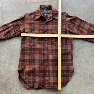 May include: A long-sleeve button-up shirt in a brown and red plaid pattern with two chest pockets and a quilted collar lining. A wooden yardstick is placed over the shirt, showing a length of 28 inches and a chest width of 21 inches.