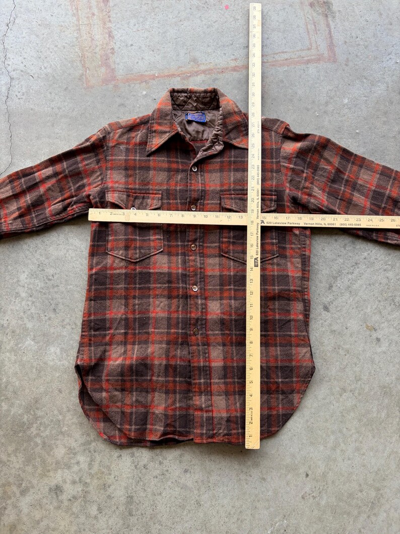 May include: A long-sleeve button-up shirt in a brown and red plaid pattern with two chest pockets and a quilted collar lining. A wooden yardstick is placed over the shirt, showing a length of 28 inches and a chest width of 21 inches.