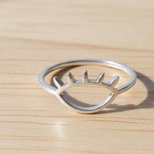 Può includere: Un anello color argento con un design a occhio aperto. L'anello presenta una linea di ciglia inferiore curva e tre ciglia dritte. L'anello è appoggiato su una superficie di legno marrone chiaro.