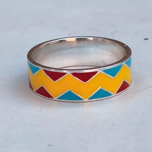 Può includere: Un anello d'argento con un design geometrico colorato. L'anello presenta una fascia di zigzag gialli, rossi e blu. Un gioiello audace e accattivante.