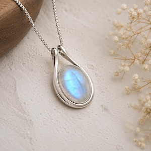 May include: A silver pendant necklace featuring an oval-shaped moonstone with a blue iridescent sheen. The stone is set in a smooth, polished silver frame that tapers upward to connect to a delicate silver box chain, creating a teardrop shape.