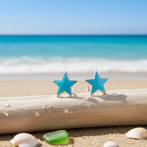 Peut inclure: Deux boucles d'oreilles en forme d'étoile avec un centre en émail turquoise vif et une bordure argentée. Les boucles d'oreilles sont présentées sur un morceau de bois flotté sur une plage de sable, avec l'océan en arrière-plan.