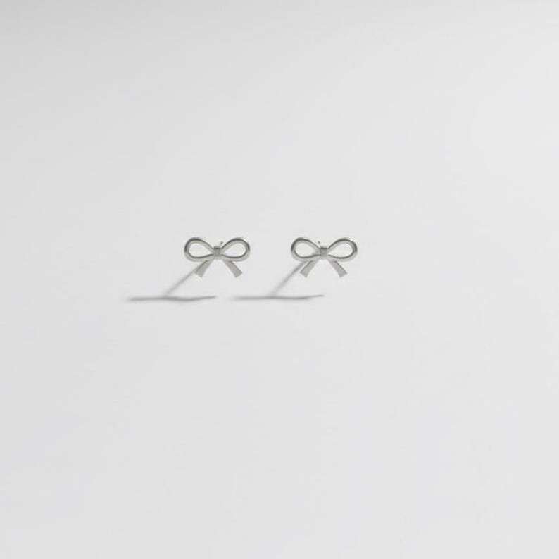 Pu&ograve; includere: Un paio di orecchini a bottone argentati a forma di fiocco. Gli orecchini sono piccoli e delicati, con un design semplice ed elegante. I fiocchi sono simmetrici e proiettano ombre sulla superficie bianca.