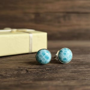 Puede incluir: Un par de pendientes de botón redondos plateados con una piedra azul claro estampada. Los pendientes están sobre una superficie de madera oscura, con una caja de regalo amarilla al fondo.
