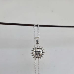 May include: A silver-toned metal necklace featuring a circular pendant with a detailed embossed design of a sun and a crescent moon face. The pendant is suspended from a delicate silver-toned chain, showcasing a polished finish with dark accents.