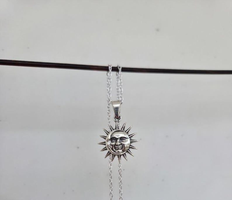 May include: A silver-toned metal necklace featuring a circular pendant with a detailed embossed design of a sun and a crescent moon face. The pendant is suspended from a delicate silver-toned chain, showcasing a polished finish with dark accents.