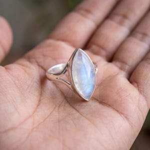 May include: A silver ring featuring a marquise-cut moonstone with a translucent, iridescent blue sheen. The band is crafted from polished silver and splits into two distinct sections as it connects to the central gemstone setting.