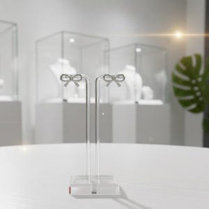 Pu&ograve; includere: Espositore per orecchini in acrilico trasparente con due montanti verticali. Ogni montante ha una parte superiore a forma di fiocco. L'espositore &egrave; su una superficie bianca, con vetrine di gioielli sullo sfondo.