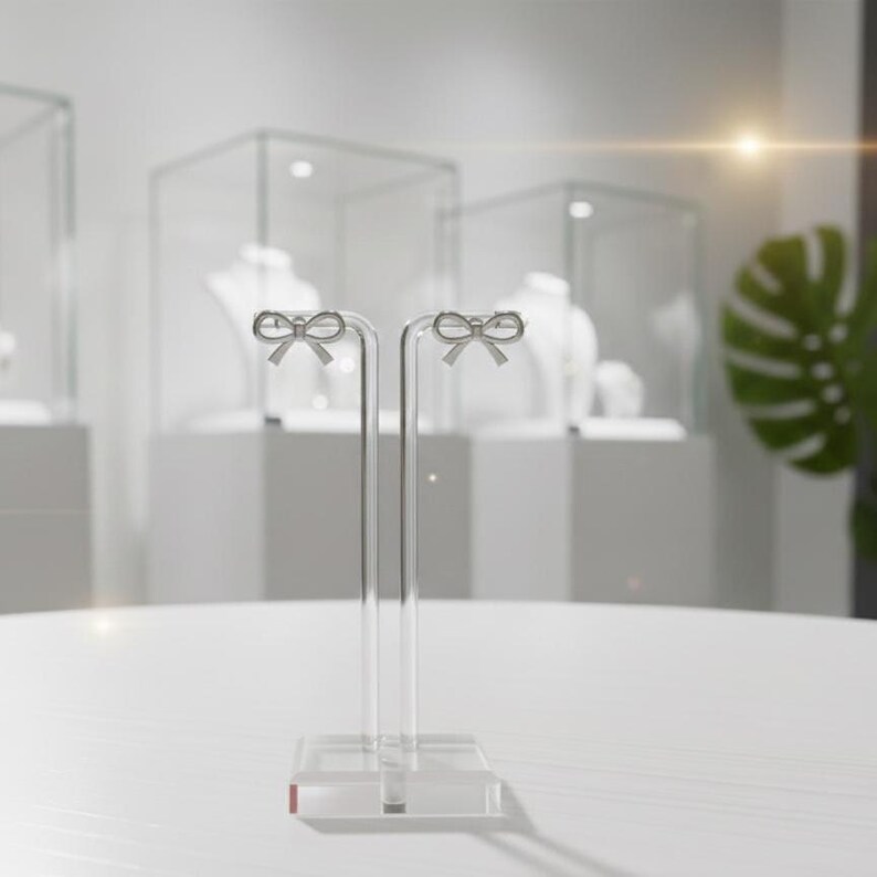 Pu&ograve; includere: Espositore per orecchini in acrilico trasparente con due montanti verticali. Ogni montante ha una parte superiore a forma di fiocco. L'espositore &egrave; su una superficie bianca, con vetrine di gioielli sullo sfondo.