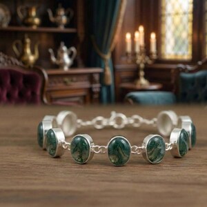 May include: A silver bracelet featuring oval-shaped green gemstones. The gemstones are set in individual silver bezels and connected by a delicate chain. The bracelet is displayed on a wooden surface.