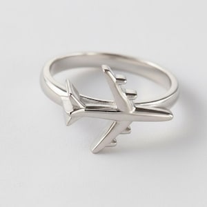 Peut inclure: Une bague argentée ornée d'un avion détaillé. La bague a une finition lisse et polie et une bande classique. L'avion a des ailes, une queue et un fuselage, créant un effet tridimensionnel. La bague est posée sur un fond blanc.
