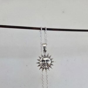 May include: A silver-toned metal necklace featuring a circular pendant with a detailed embossed face of a sun and a crescent moon. The pendant is surrounded by radiating sun rays and is suspended from a delicate silver-toned link chain.