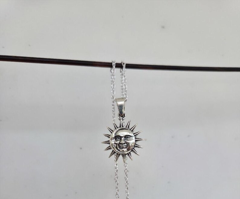 May include: A silver-toned metal necklace featuring a circular pendant with a detailed embossed face of a sun and a crescent moon. The pendant is surrounded by radiating sun rays and is suspended from a delicate silver-toned link chain.