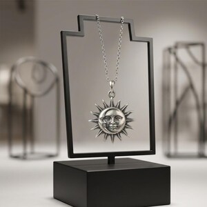 May include: A silver-toned necklace featuring a circular pendant with a sun and crescent moon face design. The pendant hangs from a delicate chain, suspended on a black rectangular display stand with a solid base, showcasing the metal craftsmanship.
