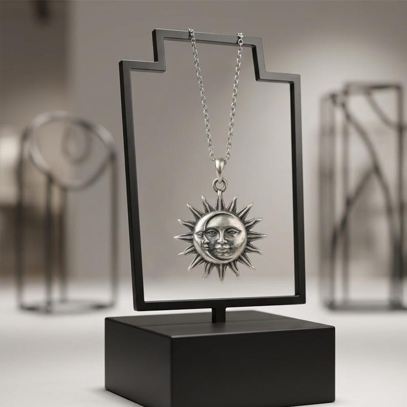 May include: A silver-toned necklace featuring a circular pendant with a sun and crescent moon face design. The pendant hangs from a delicate chain, suspended on a black rectangular display stand with a solid base, showcasing the metal craftsmanship.