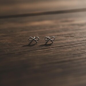 Pu&ograve; includere: Un paio di piccoli orecchini a bottone argentati a forma di fiocco. Gli orecchini sono posizionati su una superficie di legno marrone scuro, evidenziando i dettagli delicati dei fiocchi. Lo sfondo &egrave; sfocato, accentuando gli orecchini.