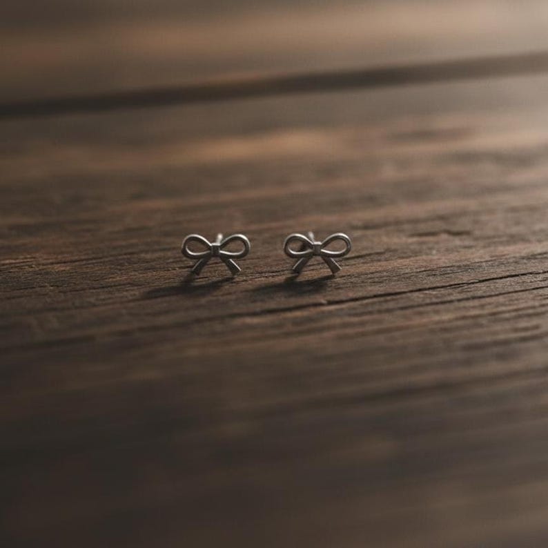 Pu&ograve; includere: Un paio di piccoli orecchini a bottone argentati a forma di fiocco. Gli orecchini sono posizionati su una superficie di legno marrone scuro, evidenziando i dettagli delicati dei fiocchi. Lo sfondo &egrave; sfocato, accentuando gli orecchini.