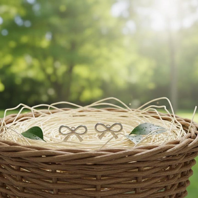 Pu&ograve; includere: Un paio di orecchini argentati a forma di fiocco in un cestino di vimini pieno di paglia. Gli orecchini hanno un design delicato ed elegante. Lo sfondo &egrave; una vista sfocata di verde, che suggerisce un ambiente naturale.