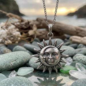May include: A silver-toned metal pendant necklace featuring a celestial design with a sun and crescent moon face. The pendant is suspended from a matching silver-toned chain, showcasing intricate facial features and radiating sun rays.