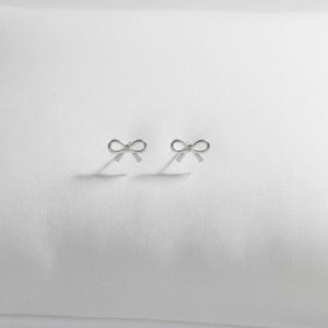 Pu&ograve; includere: Un paio di piccoli orecchini a bottone argentati a forma di fiocco. Gli orecchini sono esposti su una superficie bianca, proiettando ombre sottili. I fiocchi sono delicati e simmetrici, suggerendo un design di gioielli minimalista.