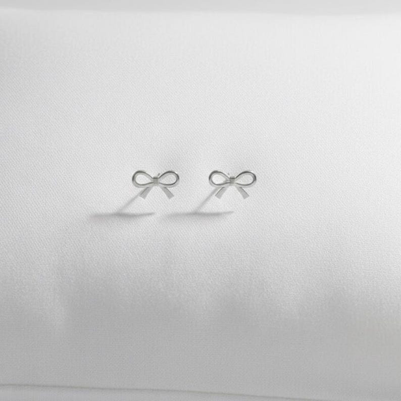 Pu&ograve; includere: Un paio di piccoli orecchini a bottone argentati a forma di fiocco. Gli orecchini sono esposti su una superficie bianca, proiettando ombre sottili. I fiocchi sono delicati e simmetrici, suggerendo un design di gioielli minimalista.