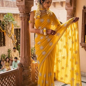 Puede incluir: Una mujer con un sari amarillo vibrante con intrincados bordados plateados. El sari está elegantemente drapeado, mostrando su tejido fluido. Está adornada con joyas florales, incluyendo un collar, aretes y brazaletes, creando un look festivo y elegante.