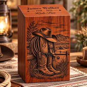 May include: A rectangular wooden cremation urn with a warm brown finish. The top is engraved with the text James Walker, 1948-2026, and Ride Free. The front features a detailed laser-engraved scene of a cowboy hat, boots, and a lasso.