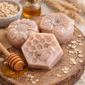 May include: Three handmade soaps with floral and honeycomb designs rest on a wooden surface. A wooden honey dipper sits in a pool of honey. Oats, honey, and a small bottle of oil are also in the image. The soaps are light brown.