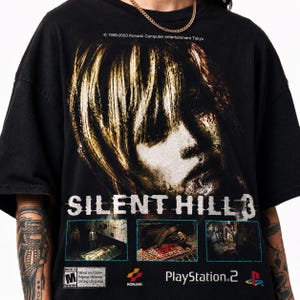 Può includere: Una t-shirt nera con una grande stampa grafica della copertina del videogioco Silent Hill 3. Il design include il titolo del gioco, tre piccoli riquadri di scene, una riga di copyright Konami, un riquadro di classificazione ESRB M e il logo PlayStation 2.